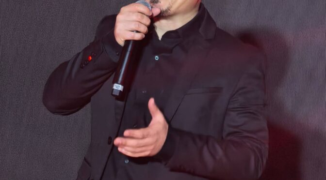 Kuya Manzano on stage holding a microphone, wearing a black suit